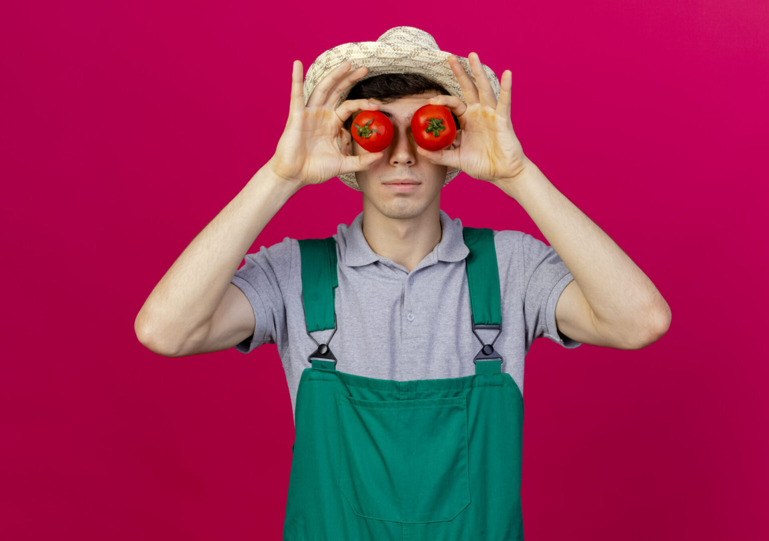 confident young male gardener wearing gardening hat covers eyes with tomatoes isolated on pink background with copy space